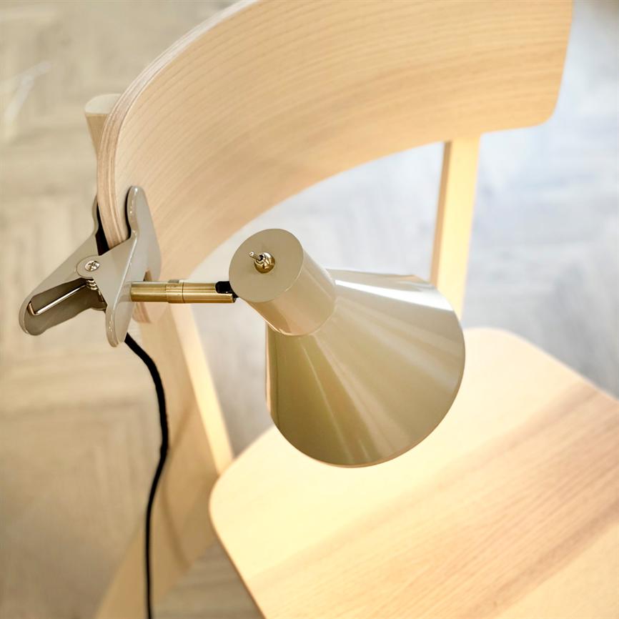Klemme-lampe fastgjort til ryglæn, vinklet ned over stolens sæde; beige-metallisk skærm og sort ledning, placeret på en lys træstol foran lyst parketgulv.