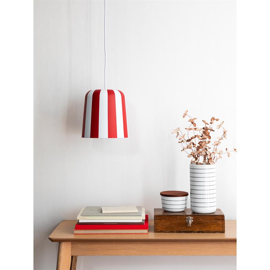 Rød- og hvidstribet pendel hænger over et lyst træbord; på bordet ligger en stak bøger ved siden af en trææske og en hvid, stribet vase med tørrede blade, hvid væg bagved.