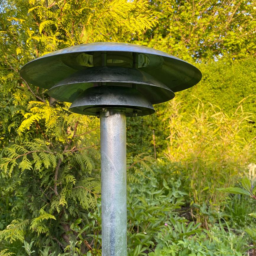 En rund, lagdelt topmonteret havelampe står på en slank metalstang blandt solbelyste buske og grønne planter i en frodig have.