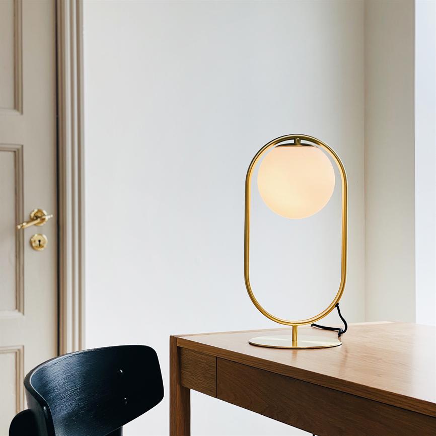 En guldfarvet, ovalt rammebordlampe med rund, varmglødende skærm; den står tændt på et træbord ved en sort stol foran en hvid væg og en dør med messinghåndtag.