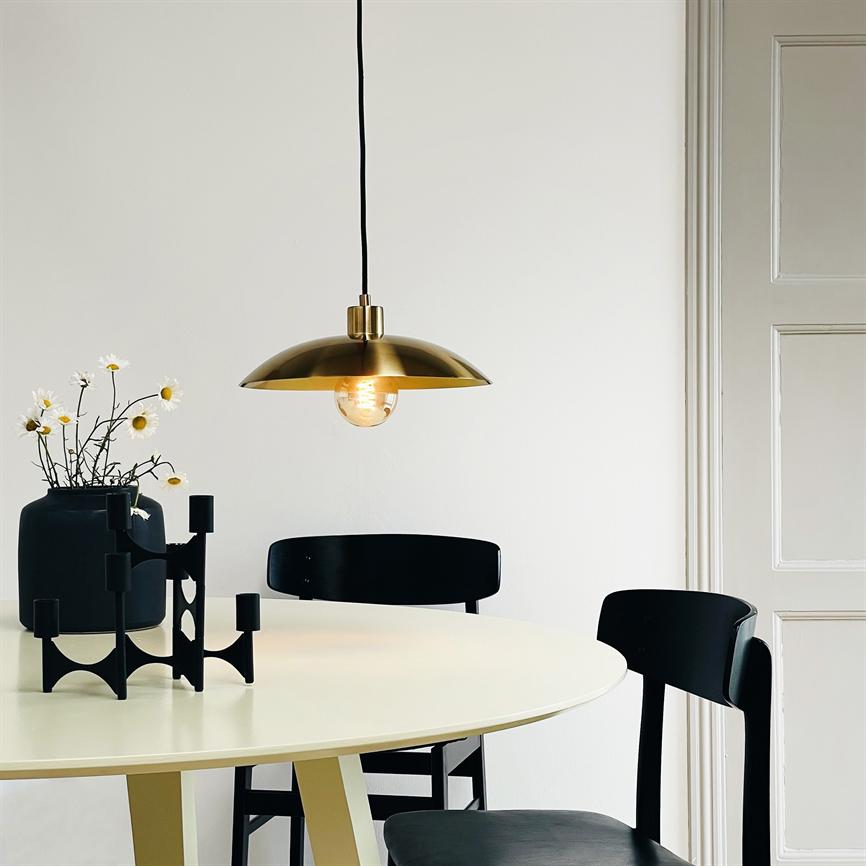 En messingpendel hænger og kaster varmt lys over et rundt hvidt bord; omkring står sorte stole, en sort vase med marguritter og et geometrisk sort lysestage mod hvid væg.