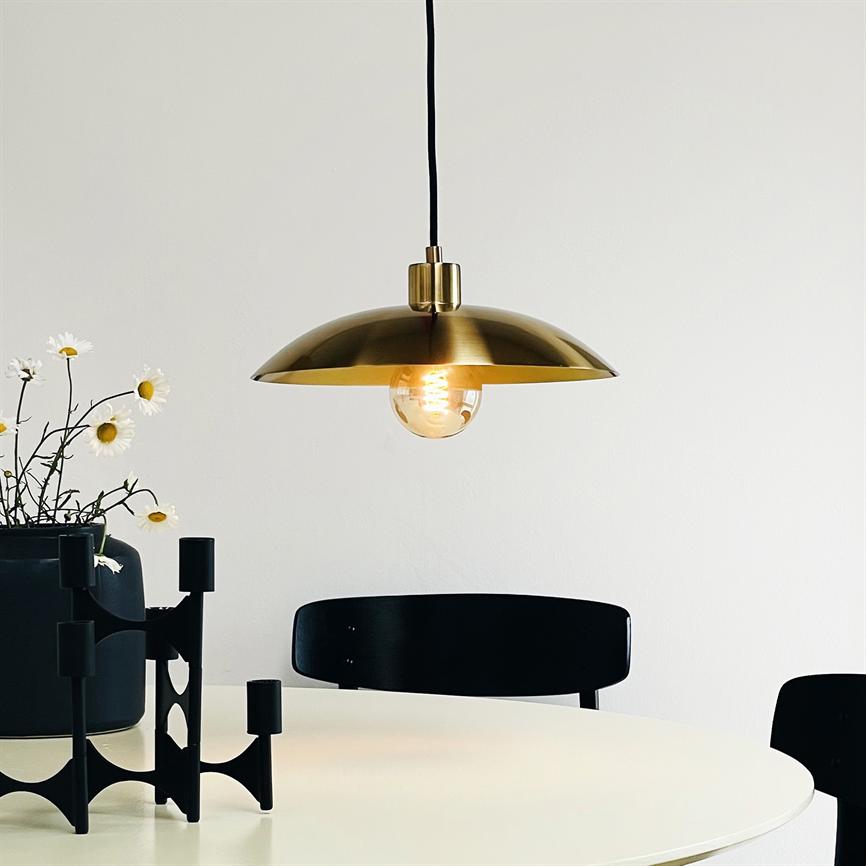 En messingpendel lyser med en synlig pære, hænger over et rundt hvidt bord; omkring står sorte stole, en sort lysestage og en vase med hvide margueritter mod en lys væg.