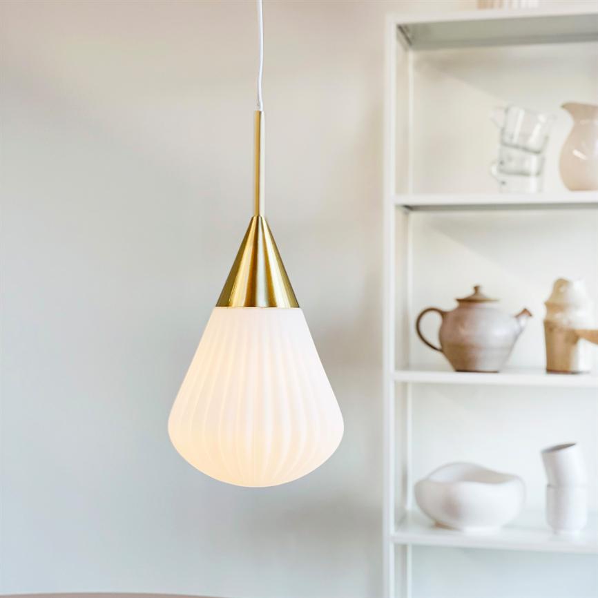 En hvid, rippet pendel med guldfarvet top hænger og afgiver blødt lys foran en hvid reol med keramikkander, skåle og krus.