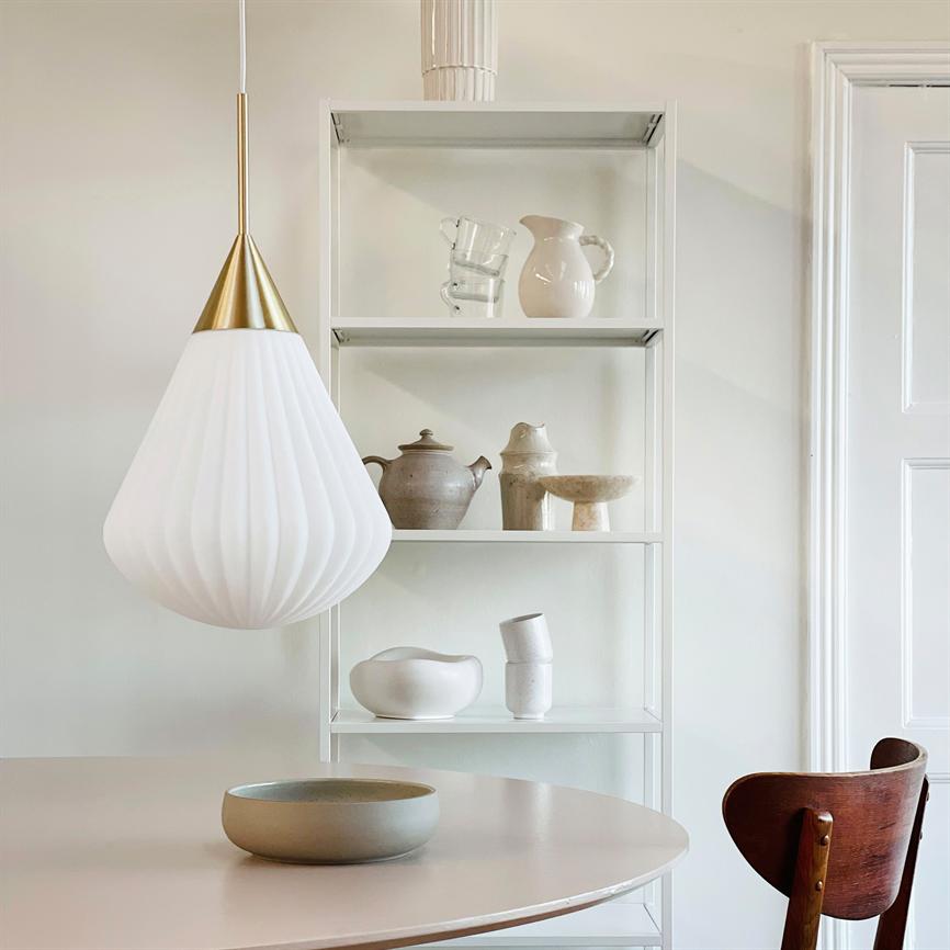 Hængende hvid dråbeformet lampe svæver over et rundt lyst spisebord; i baggrunden står en åben hvid reol med keramiske vaser og en mørk træstol.