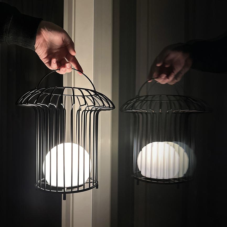 Metal-lanterne med rund hvid pære holdes af en hånd; den udsender varmt lys, og dens spejlbillede reflekteres i en mørk vinduesrude bagved.