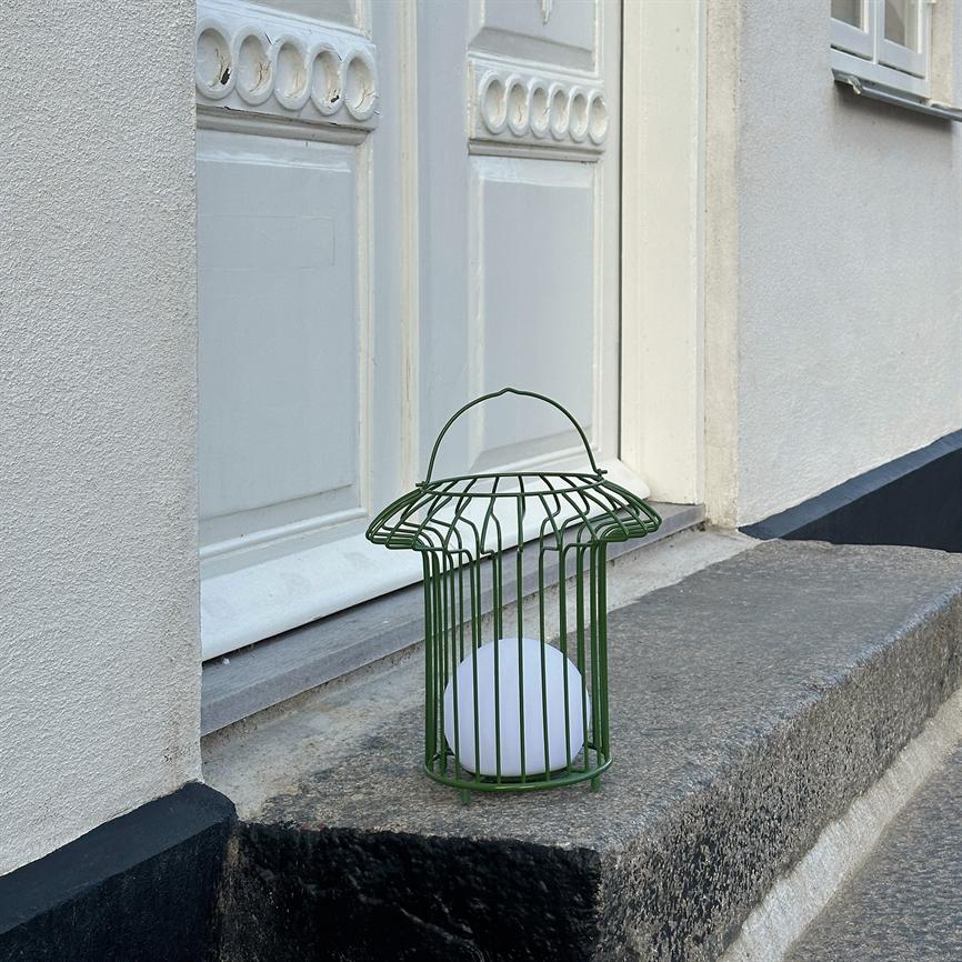 En grøn metallygte med en hvid kugle indeni står på en grå granittrappe foran en hvid dør med runde dekorative udskæringer; hvid facade og et vindue ses til højre.