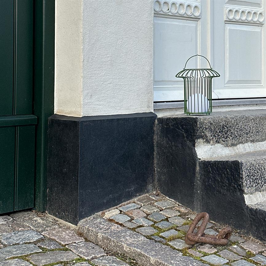 En grøn metallygte står på dørtrinnet og indeholder et hvidt lys; den står ved en hvid dør med sort sokkel, grå trapper og brostensbelagt fortov med et rustent anker.