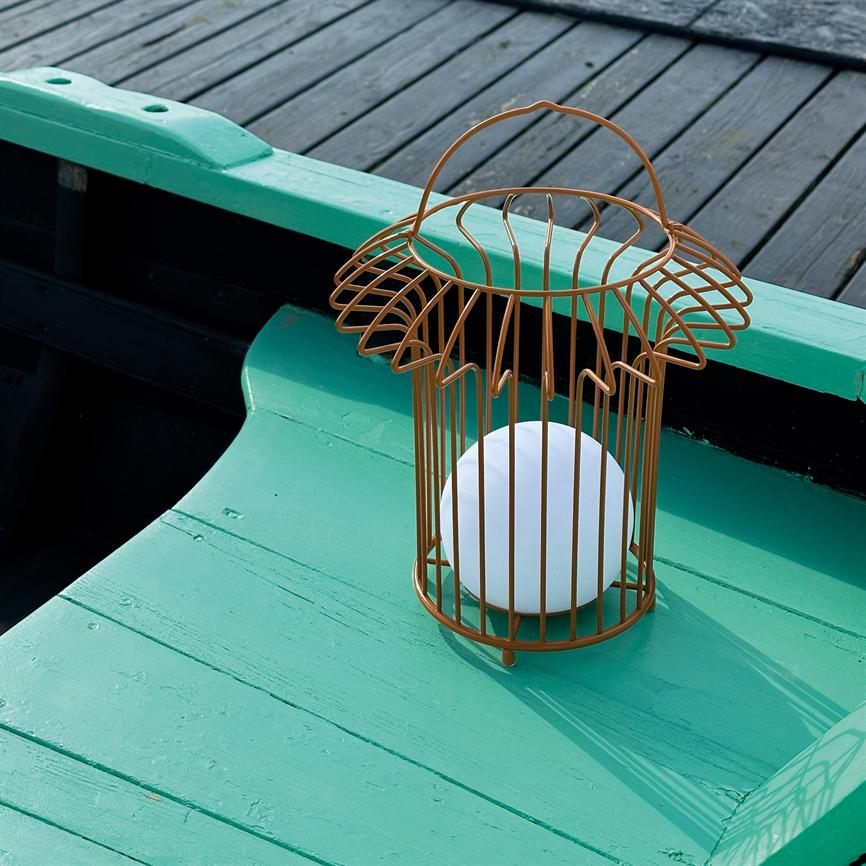 En kobberfarvet trådkurv-lanterne med en hvid kugle står på en turkismalet træplanke i en båd; mørke træbrobrædder ses i baggrunden.