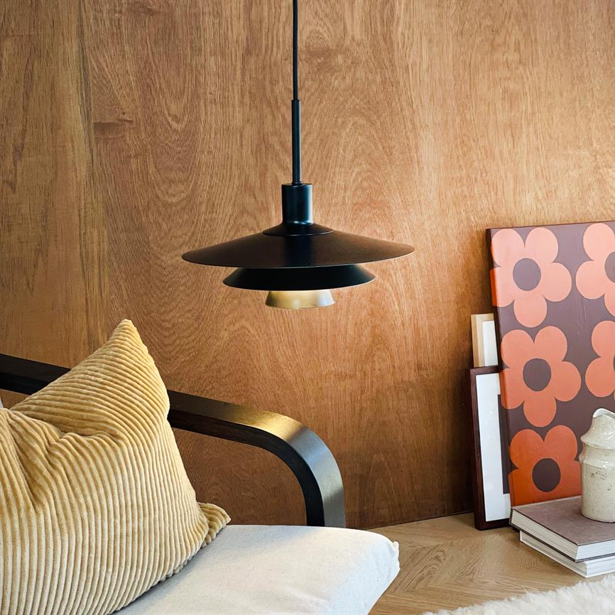 Sort pendellampe hænger og kaster blødt lys over en lænestol med beige ribbet pude; træbeklædt væg bagved, stak bøger, keramikkande og et orange blomstermotiv lænet mod væggen.