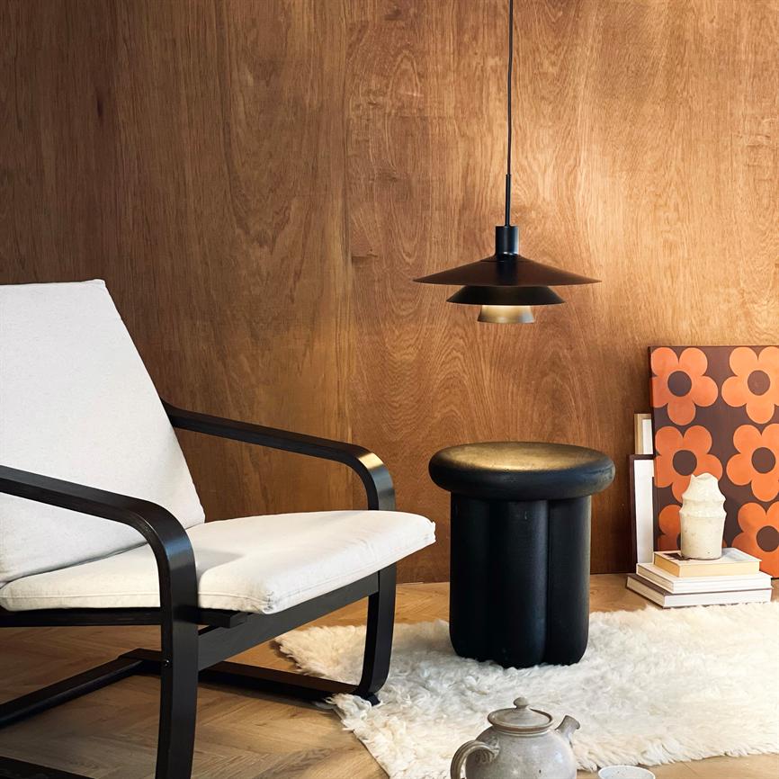 Lænestol med lyst betræk står ved et rundt sort skammel; skammelen står under en hængende sort lampe på et hvidt pels-tæppe foran en varm træbeklædt væg med bøger og blomsterplakat.