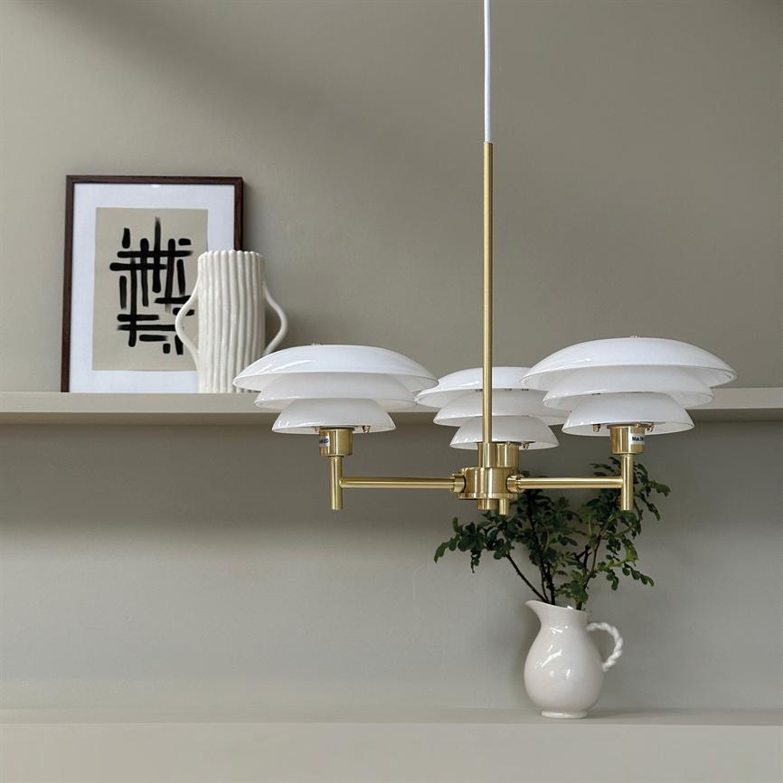 Messingpendel med seks hvide skærme hænger over en hylde; på hylden en hvid kande med grønt kvist og en indrammet abstrakt plakat mod beige væg. Ingen læsbar tekst.