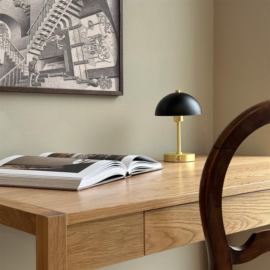 En åben bog ligger på et lyst træskrivebord; en lille sort‑og‑guld bordlampe står slukket ved siden af, mens en grafisk sort‑hvid plakat hænger på væggen og en træstol kaster skygge.