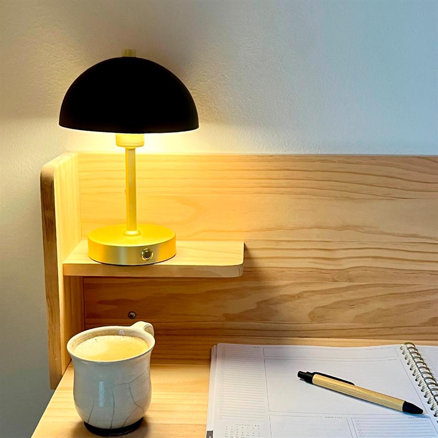 Gul bordlampe med sort kuppel lyser varmt over et lyst træskrivebord; en keramikkop med kaffe står til venstre, og en åben spiralnotebook med pen ligger til højre.