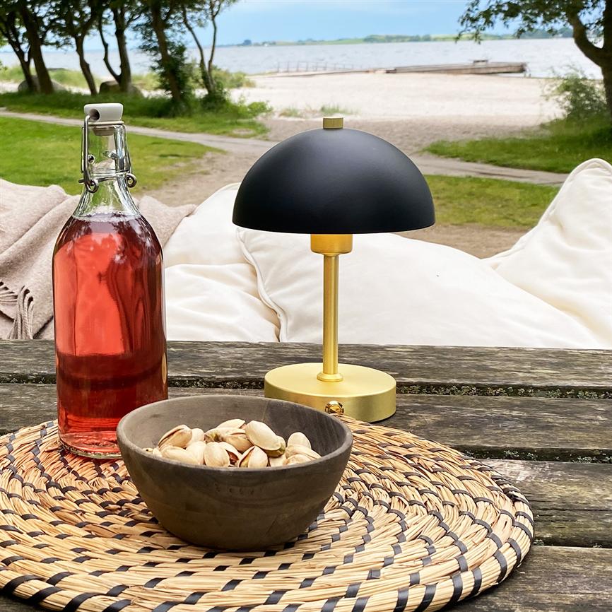 Sort-guld bordlampe står på et vejrbidt træbord; foran ligger en skål med pistacienødder på en flettet dækkeserviet, en flaske lyserød drik ved siden; strand og hav i baggrunden.