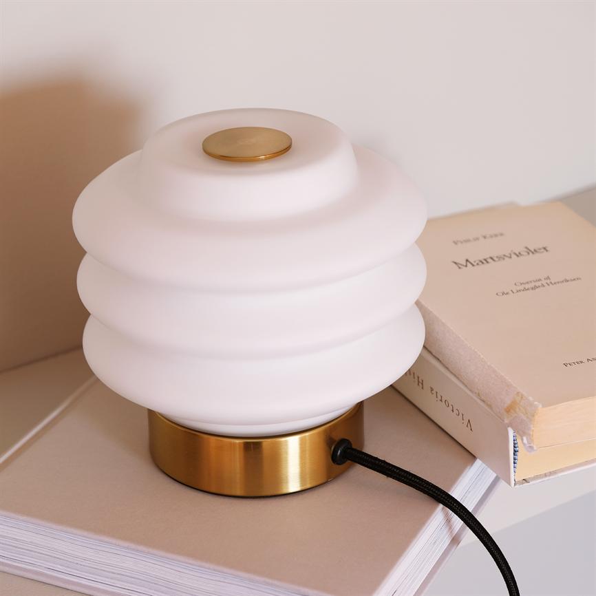 Hvid, bølget bordlampe med messingfod står slukket på en stak bøger i et lyst, minimalistisk rum. Der er tekst på en bog, men den er utydelig og ikke læselig.