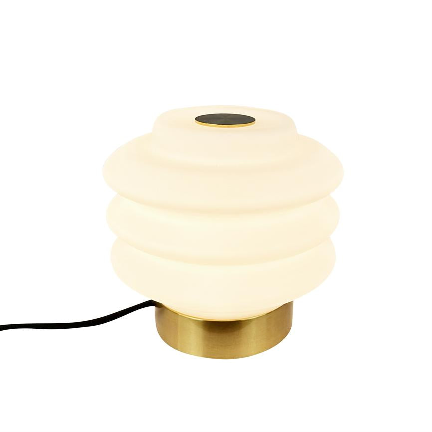 Lampe lyser blødt; rund, ribbet hvid glaskuppel på lav, børstet messingfod; sort ledning løber ud til siden på neutral, hvid baggrund.