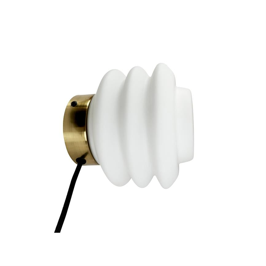 Væglampe med hvid bølget glasskærm monteret på en rund messingbase; en sort stofledning hænger nedad, vist i profil mod en hvid baggrund.