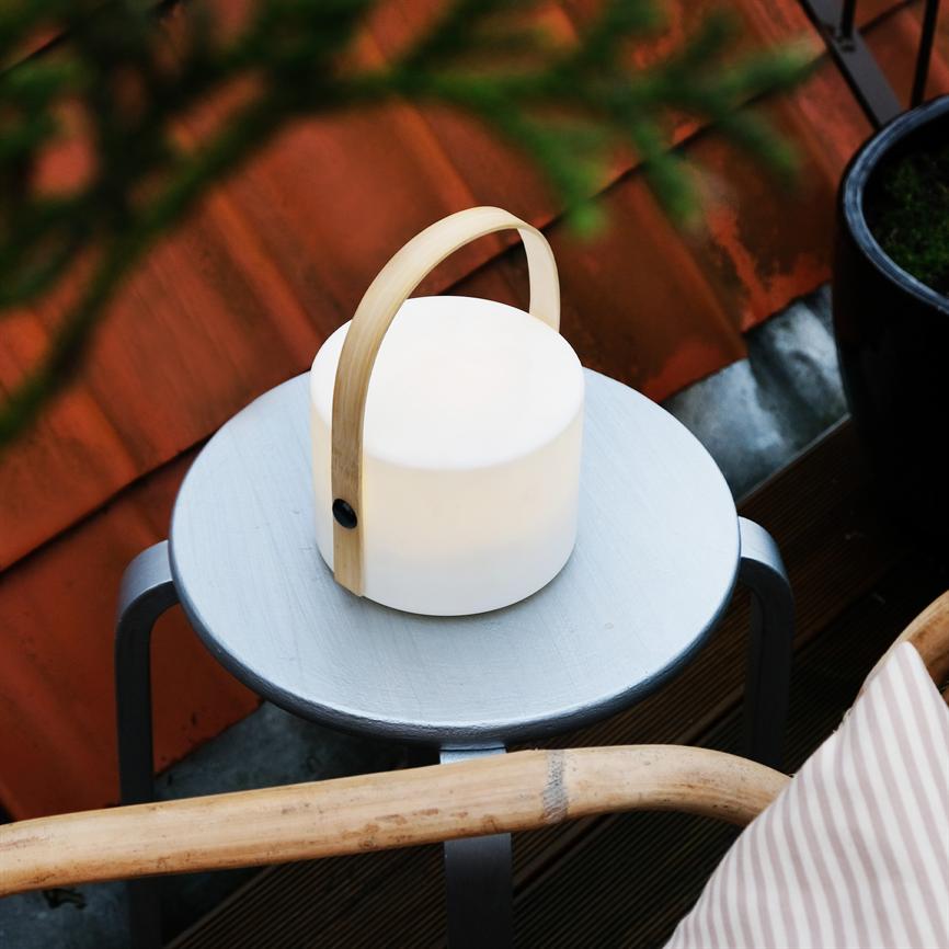 En bærbar, hvid lampe med træhåndtag står og udsender blødt lys på et rundt, lysegråt metalbord på en lille balkon med røde tagfliser, kurvestol og urtepotte.