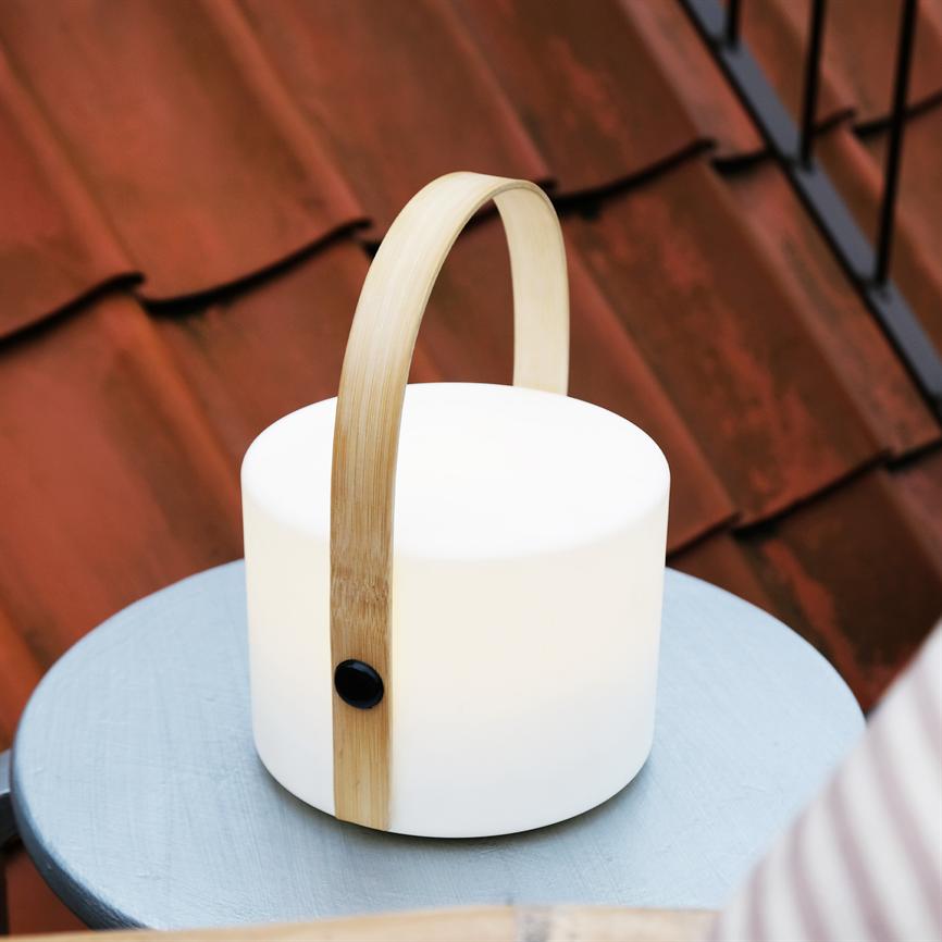 Hvid cylindrisk lampe med buet træhåndtag står på et lille blåt rundt bord. Den udsender blødt lys, placeret foran røde tagsten i et udendørs balkonmiljø.