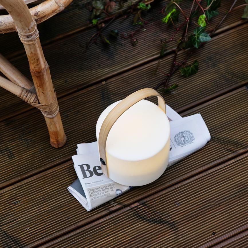 Rund bærbar lampe med bambushåndtag afgiver blødt lys, står på en sammenfoldet avis på en mørk træterrasse ved en rottingstol og klatrende efeu. Tekst: "Be"