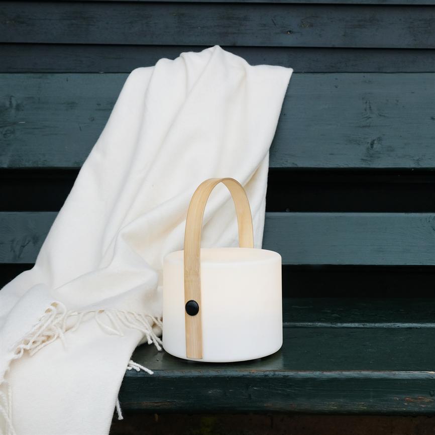 Bærbar hvid lanterne med bambushank står på en mørkegrøn træbænk og kaster blødt lys, mens et cremefarvet tæppe med frynser er smidt over ryglænet.