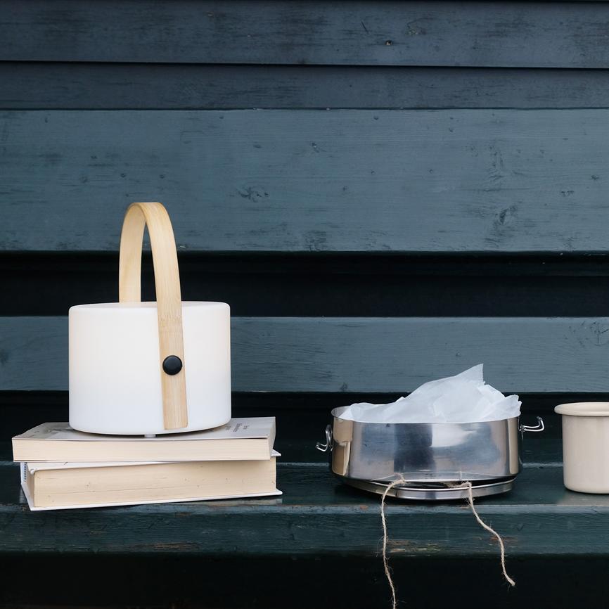 Lanterne med cylinderformet hvid skærm og træhank står på to stablede bøger; ved siden af en metalbeholder med bagepapir og en keramikkrus på en mørk bænk foran blå trævæg.