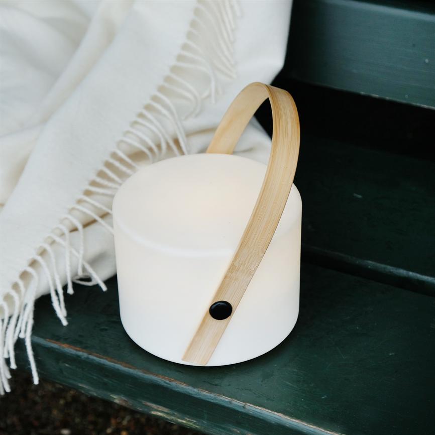 Bærbar hvid lampe med buet træhåndtag lyser blødt på en mørkegrøn træbænk ved siden af et hvidt tæppe med frynser.