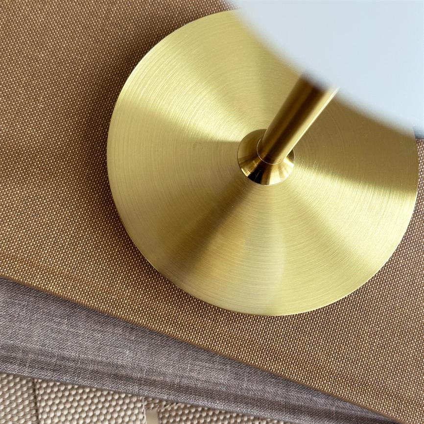 En rund messing lampefod med lodret stang hviler på lag af brune og grå tekstiler; dens skinnende, cirkulære overflade reflekterer lys på en vævet dækkeserviet og linned.