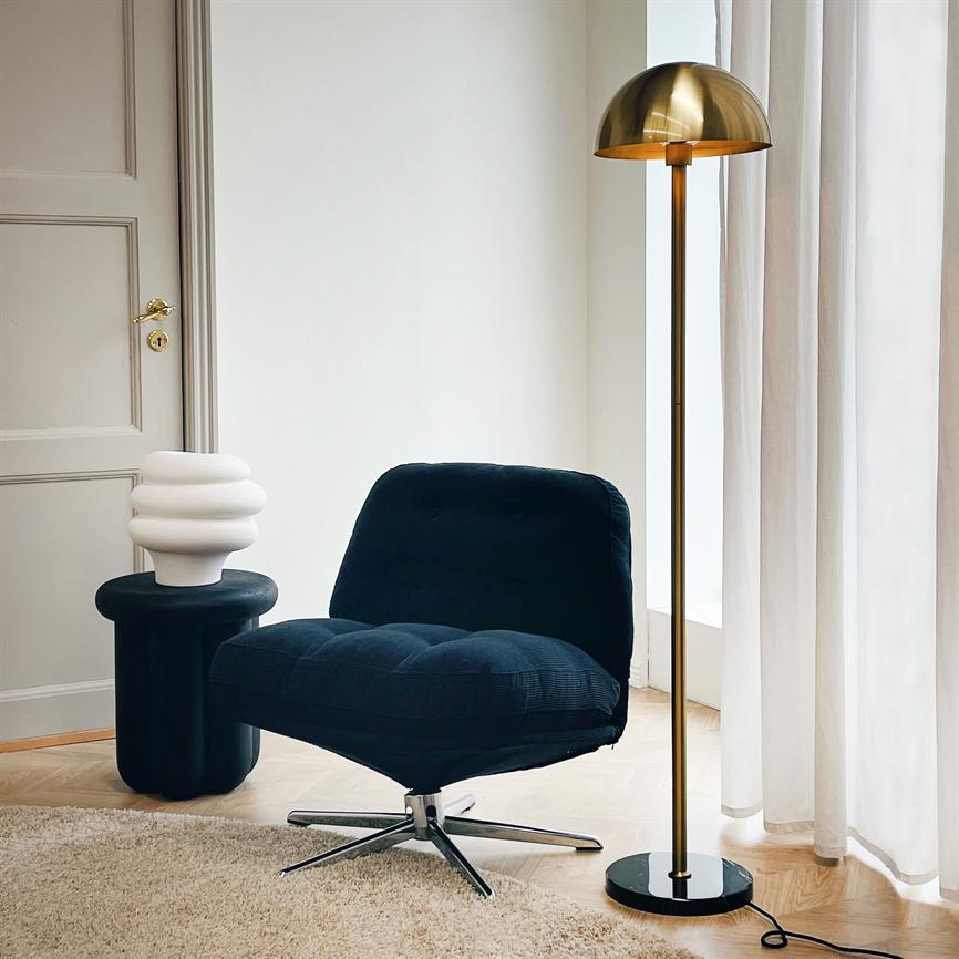 Mørk blå, polstret lænestol står på en metal drejefod ved en messing gulvlampe og et lille sort sidebord med hvid skulpturel lampe i et lyst værelse med hvide gardiner.