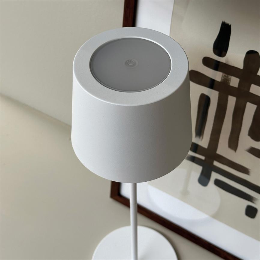 Hvid bordlampe står oprejst, top viser berøringsknap med tænd/sluk-symbol; tynd stang og rund fod på lyst bord foran et indrammet abstrakt sort-maleri.