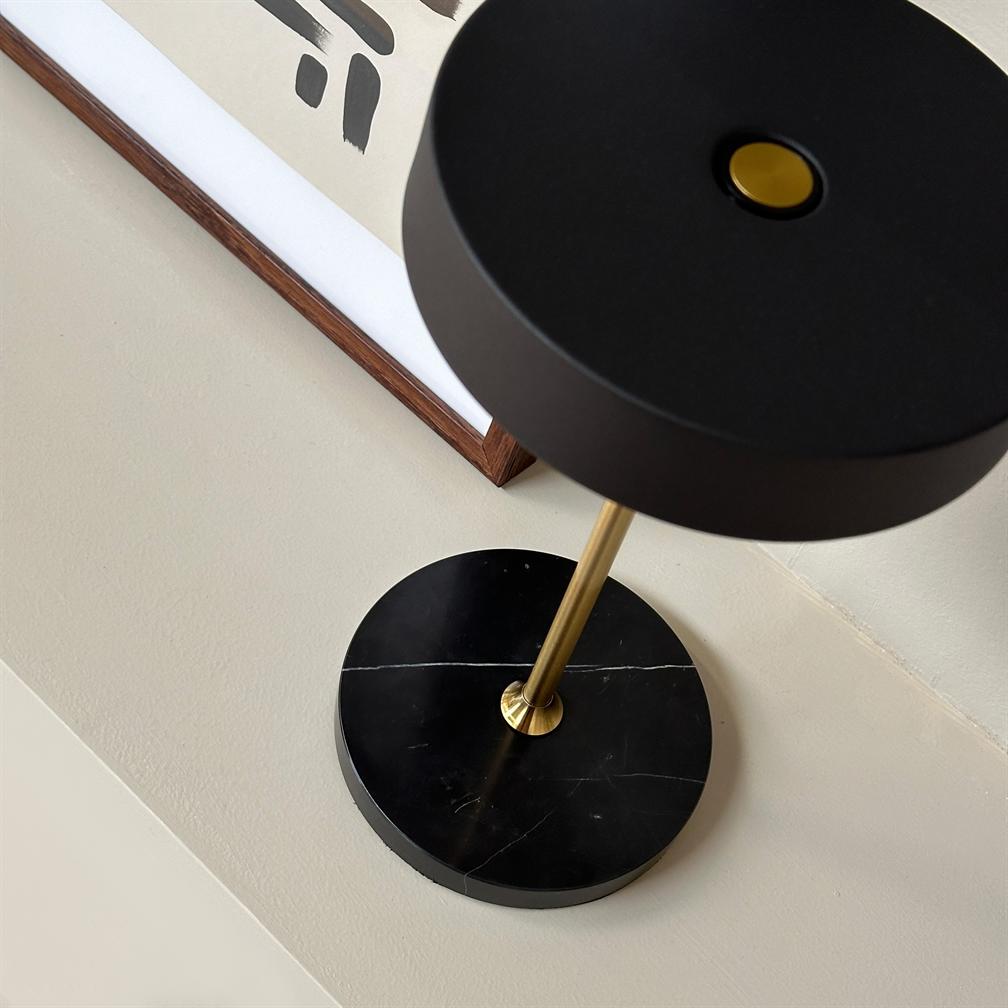 Sort bordlampe står på et beige bord; en gylden metalstang forbinder en rund sort skærm med en cirkulær sort marmorfod; bagved hænger en indrammet abstrakt plakat med sorte penselstrøg.