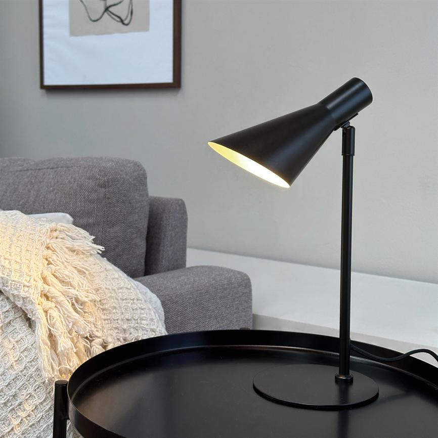 Sort metalbordlampe kaster varmt lys ned på et rundt sort sidebord ved en grå sofa med cremefarvet, textured plaide, foran en lys væg med en indrammet, minimalistisk plakat.