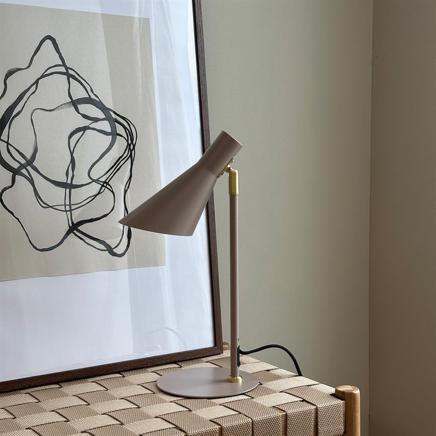 Bordlampe i beige står vinklet mod et indrammet abstrakt motiv, ikke tændt, placeret på en flettet bænk foran en lys væg i et roligt hjørne.