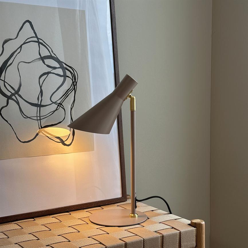 Bordlampe i mat beige kaster varmt, gyldent lys på en indrammet abstrakt linjetegning og et flettet bænkeunderlag ved en lys, neutral væg.