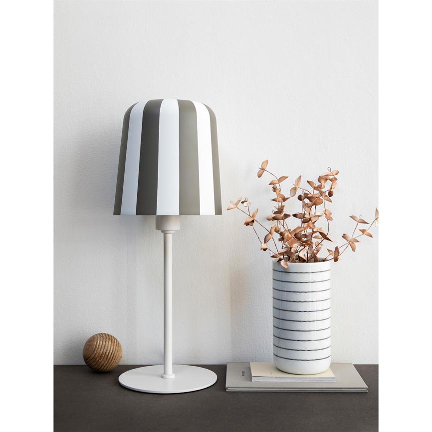 Stribet bordlampe står på en fod; ved siden af står en hvid vase med striber, fyldt med tørrede kobberfarvede blade på bøger på et mørkt bord mod en hvid væg.