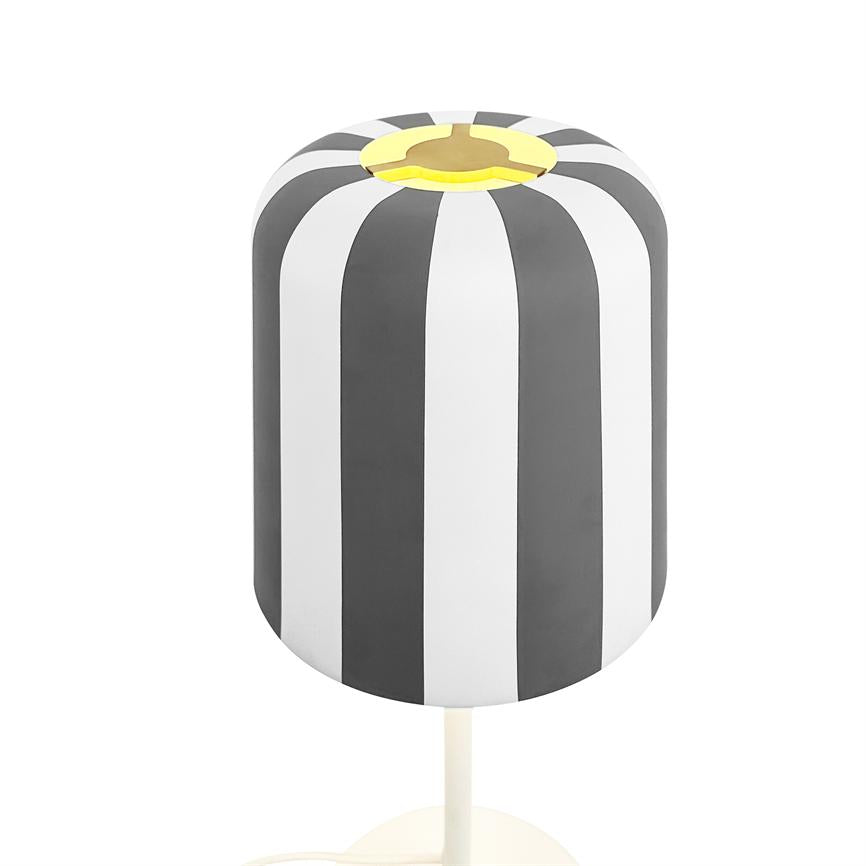 En cylindrisk bordlampe med lodrette sorte og hvide striber, gul kant i toppen, står på en hvid stolpe mod en klar, studiolignende hvid baggrund.