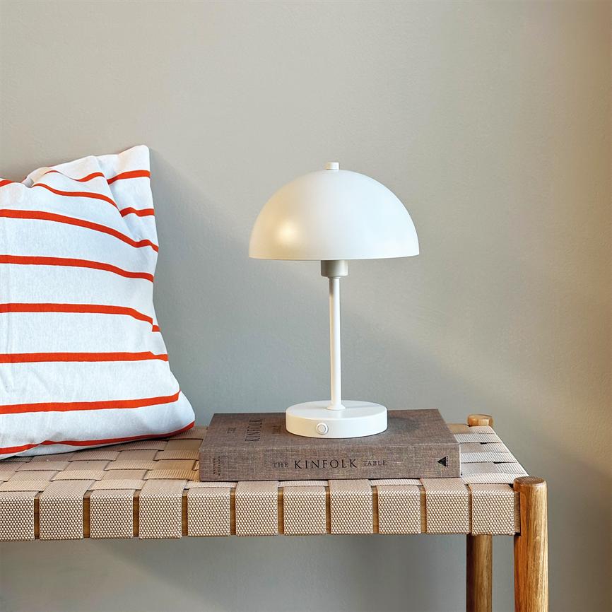 Hvid bordlampe står på en bog på en vævet træbænk ved en hvid pude med røde striber foran en beige væg. Tekst: "THE KINFOLK TABLE"