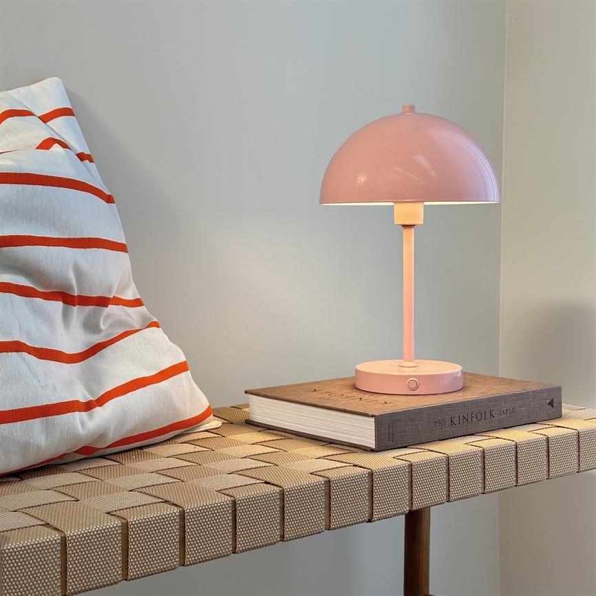 En rosa bordlampe lyser blødt på en bog med teksten THE KINFOLK TABLE, placeret på en flettet bænk ved en hvid væg og en hvid pude med orange striber.