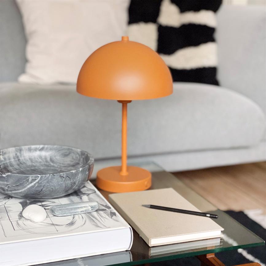 Orange bordlampe står på et sofabord af glas foran en grå sofa; ved siden af ligger en marmoreret skål ovenpå en stor bog samt en notesbog med en sort blyant.