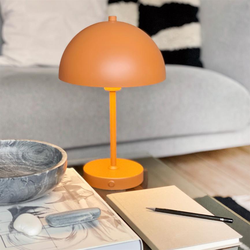 Orange bordlampe står på et glasbord; den lyser svagt over en notesbog og en HB-blyant. I baggrunden ses en grå sofa og en sort-hvid dekorativ pude.