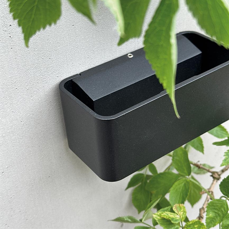 Sort, rektangulært vægmonteret beholder sidder på en lysegrå væg, delvist skjult af grønne blade fra en plante, med afrundede hjørner og et åbent toprum.