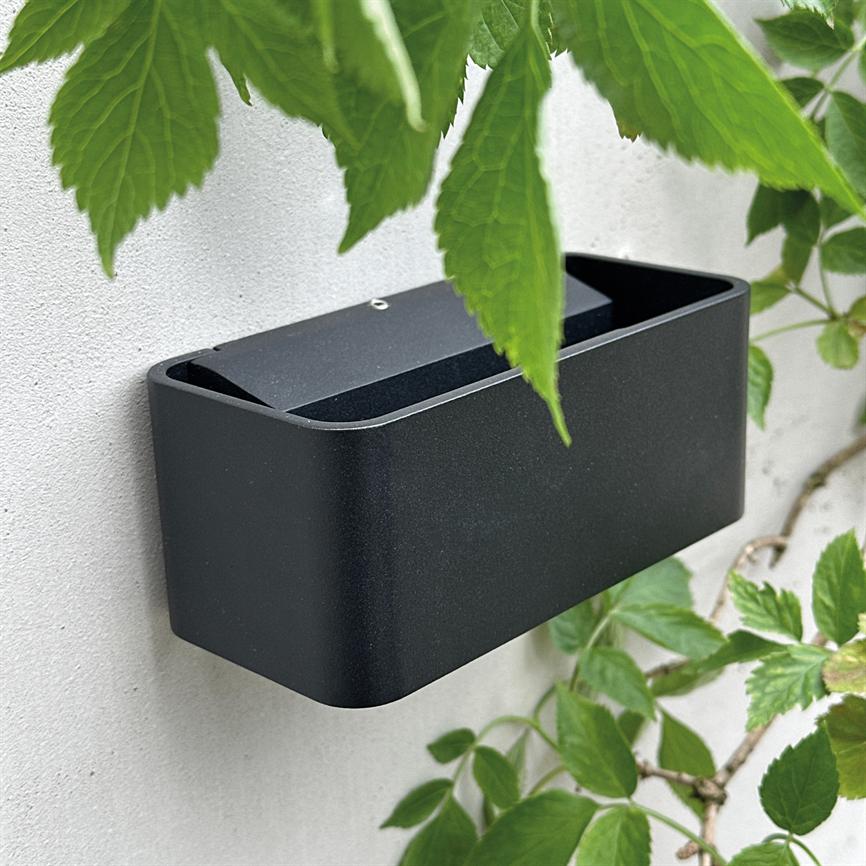 En sort, vægmonteret plantekasse hænger på en hvid mur; den er tom med afrundede hjørner og omgivet af grønne klatreblade.