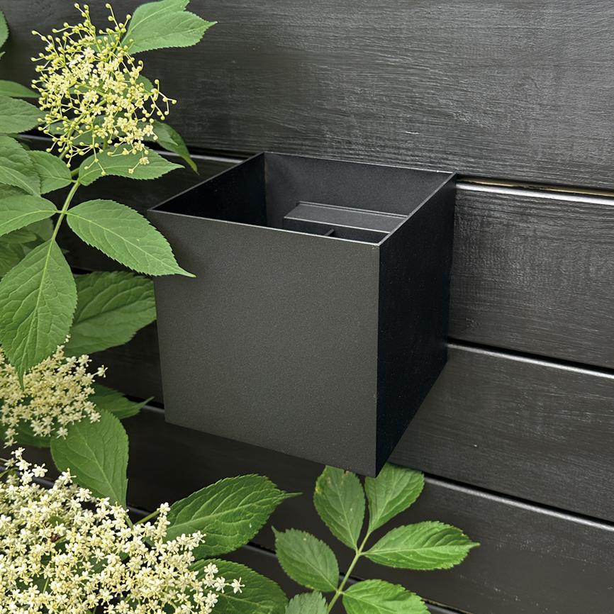 Sort firkantet metalboks monteret på et mørkt, horisontalt træhegn; omgivet af grønne blade og klaselignende små hvide blomster.