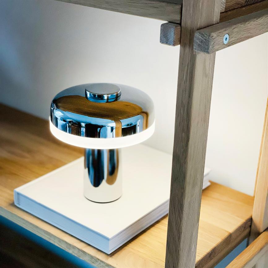 Kromfarvet svampelampe udsender hvidt ringelys, står på en lukket hvid bog på en lys træhylde under en rå egetræsreol i et roligt, moderne interiør.