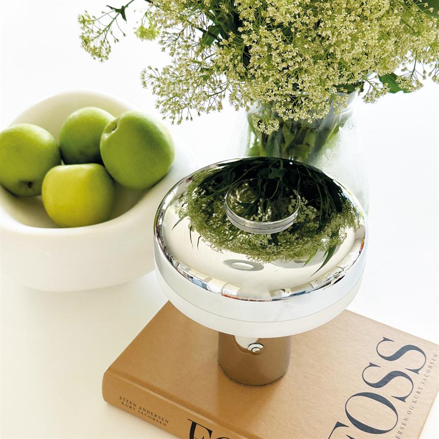 Rund, poleret metaldome står på en bog og reflekterer en vase med små hvide blomster; en skål med fire grønne æbler står ved siden af på et lyst bord. Tekst på bogen: "OSS".