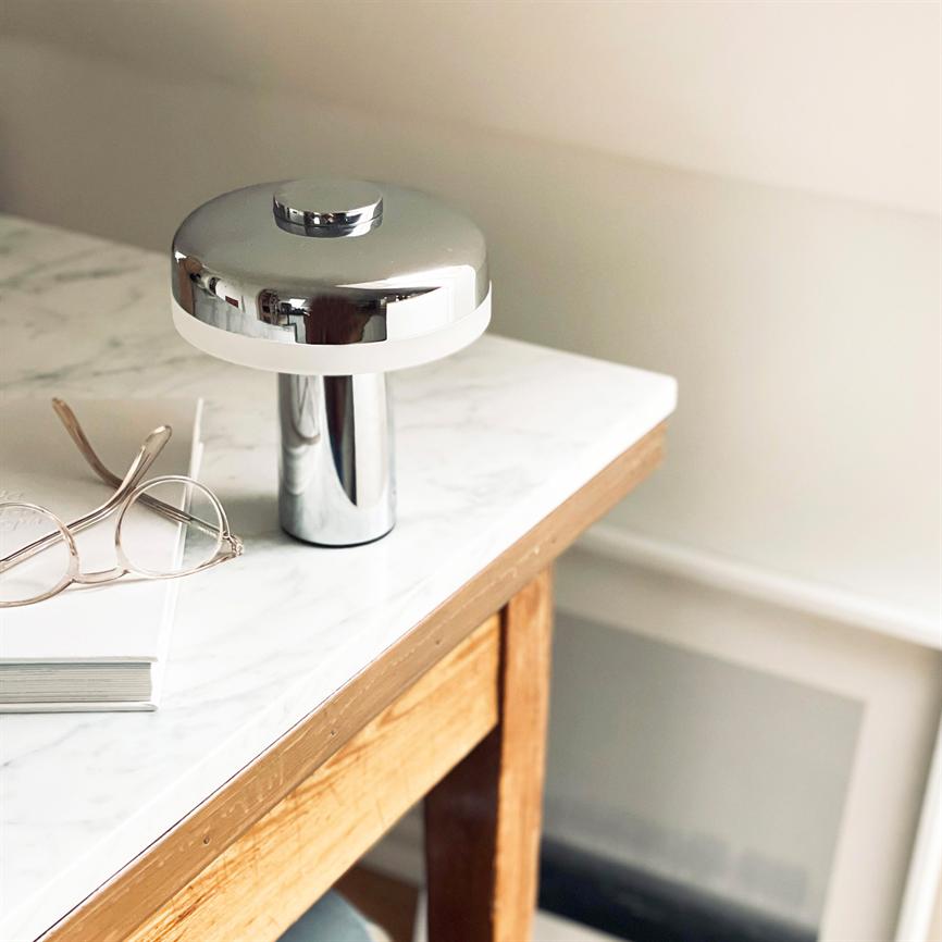 En krom bordlampe står slukket på en hvid marmorplade på et træskrivebord, flankeret af briller og en bog, i et lyst og neutralt indendørs rum.