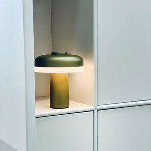 Bordlampe står tændt på en hylde i et hvidt skabsniche; olivengrøn rund skærm og cylinderformet fod, blødt varmt lys kaster skygger på bagvæggen.