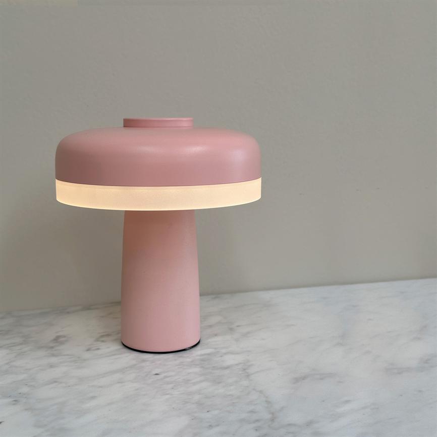 Rosa bordlampe med cylindrisk fod udsender blødt, varmt lys fra en lysrand og står på en hvid marmoroverflade foran en lysegrå væg.