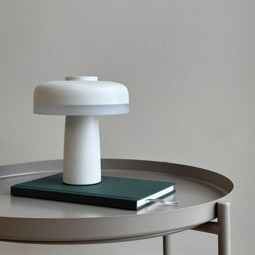 Bordlampe i hvid form som en svamp står på en mørkegrøn indbundet notesbog med bogmærkebånd, der ligger midt på et rundt gråt metalbord foran en lysgrå væg.