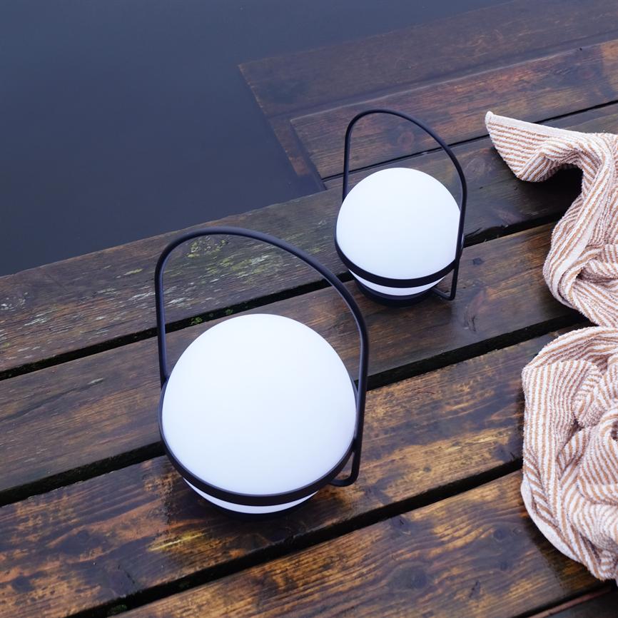 To runde lanterner med sorte håndtag hviler på en våd træbro ved mørkt vand; et stribet håndklæde ligger i nærheden.